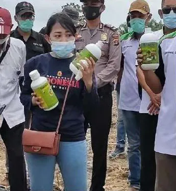Seorang perempuan bermasker mengangkat dua botol berlabel hijau di tengah kerumunan di lahan terbuka; pria di sebelah kanan juga memegang botol serupa.