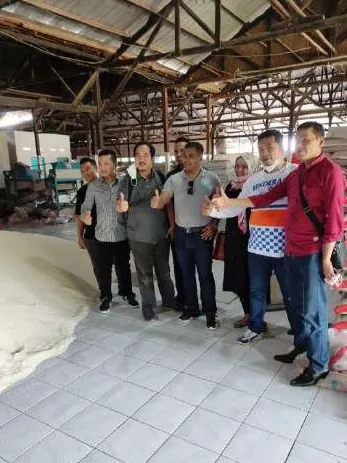 Sekelompok orang berpose sambil mengacungkan jempol di dalam gudang beratap seng dengan rangka atap terbuka, di samping hamparan bahan berwarna putih di lantai keramik.