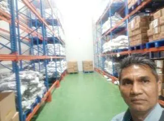 Lorong gudang dengan rak baja biru-oranye berisi karung putih dan kardus di atas lantai hijau; seorang pria berswafoto di sudut kanan bawah.