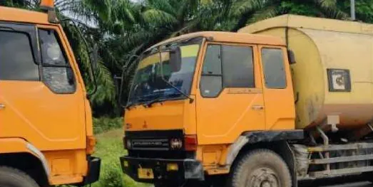 Dua truk berkabin oranye, salah satunya truk tangki, terparkir di depan rimbun pepohonan.